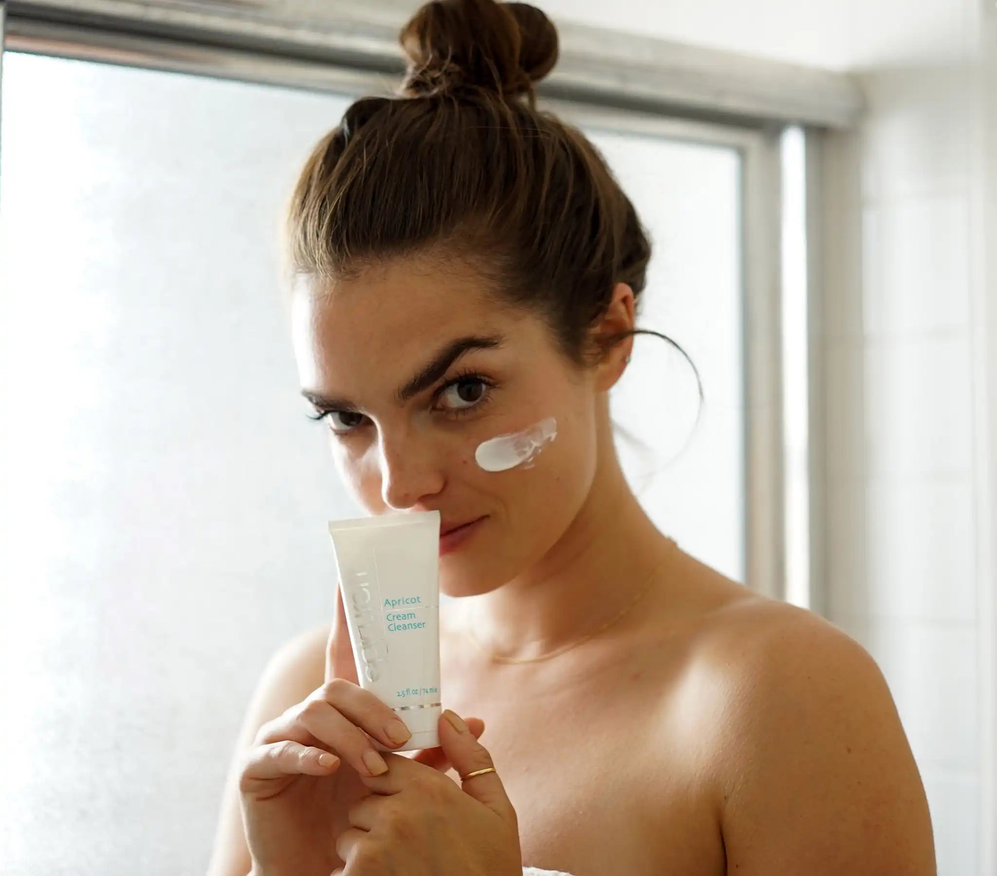 Woman applying cream to her face with a tube of cream in front of a window.