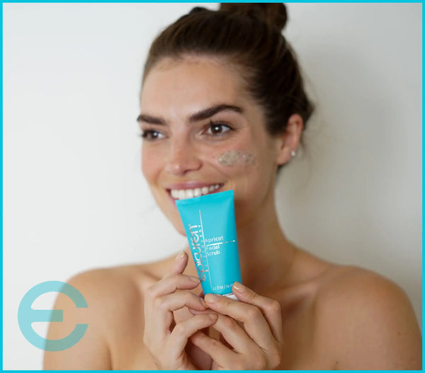 Woman smiling and holding a tube of Epicuren Apricot Facial Scrub, with some of the scrub applied to her cheek.